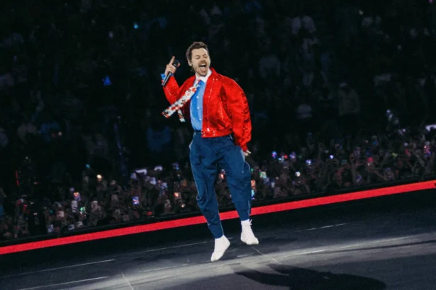 Harry Styles leaps mid-air while singing into a microphone on stage, wearing a red satin jacket, blue trousers, and white socks, with a packed crowd holding up phones behind him.