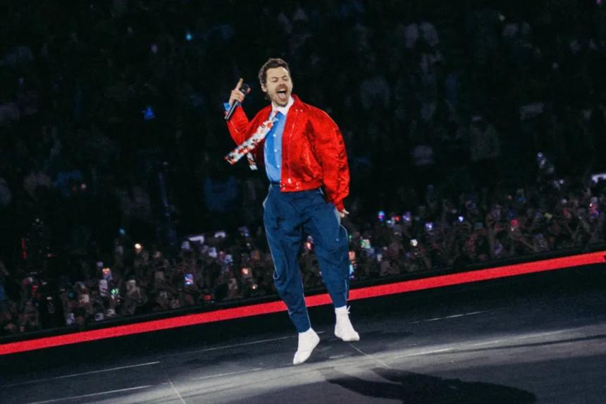 Harry Styles leaps mid-air while singing into a microphone on stage, wearing a red satin jacket, blue trousers, and white socks, with a packed crowd holding up phones behind him.
