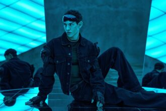Nick Hissom poses in a denim outfit with armored accents on a mirrored floor under blue neon lighting, in press art for his EP 'Hot Boy Things.'