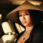 Jhené Aiko poses in warm golden light wearing a wide-brimmed conical straw hat and layered silver necklaces, with soft glittery accents beneath her eyes.