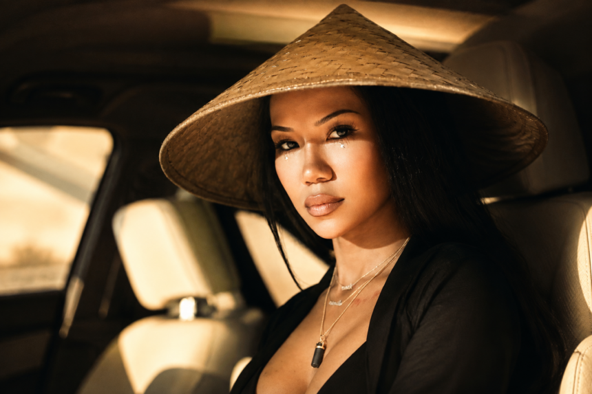 Jhené Aiko poses in warm golden light wearing a wide-brimmed conical straw hat and layered silver necklaces, with soft glittery accents beneath her eyes.