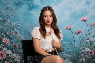 Laufey sits on a black chair holding a single gerbera daisy against a dreamy floral backdrop of pink and white blooms, wearing a white lace blouse and plaid skirt with a soft smile.