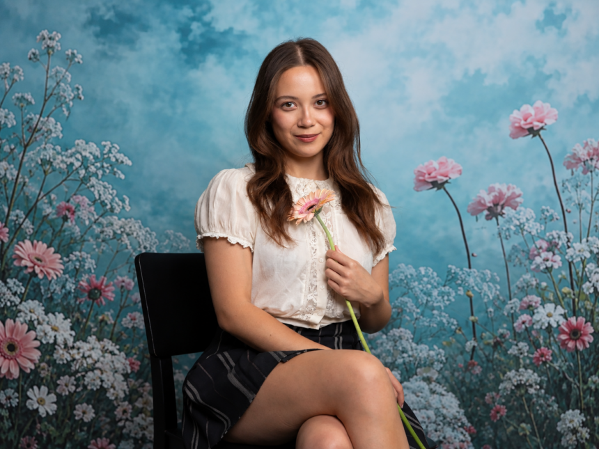 Laufey sits on a black chair holding a single gerbera daisy against a dreamy floral backdrop of pink and white blooms, wearing a white lace blouse and plaid skirt with a soft smile.