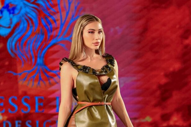 Lika O walks the runway for Jesse Lee Designs at New York Fashion Week in an olive leather cutout gown with a thin belt and strappy knee-high buckled boots, against a red backdrop bearing the designer's name.