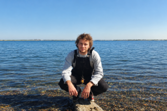 Mac DeMarco crouches on a pebbled shoreline in front of calm blue water under a clear sky, wearing a gray sweater and overalls with a keyring on his belt.