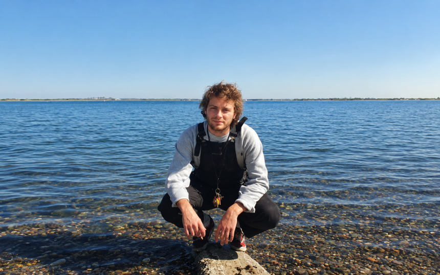 Mac DeMarco crouches on a pebbled shoreline in front of calm blue water under a clear sky, wearing a gray sweater and overalls with a keyring on his belt.