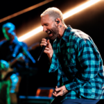 Adam Levine of Maroon 5 sings passionately into a microphone on stage in a green plaid shirt, with a bandmate playing guitar under streaking stage lights behind him.