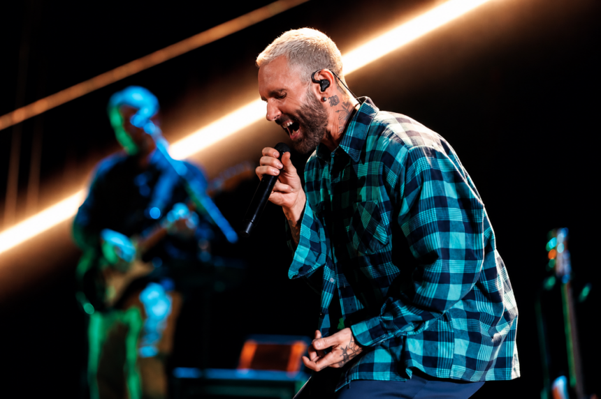 Adam Levine of Maroon 5 sings passionately into a microphone on stage in a green plaid shirt, with a bandmate playing guitar under streaking stage lights behind him.