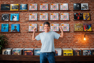 Mike D of the Beastie Boys raises both fists while standing in a record shop, in a light gray T-shirt and jeans before an exposed brick wall lined with vinyl albums.