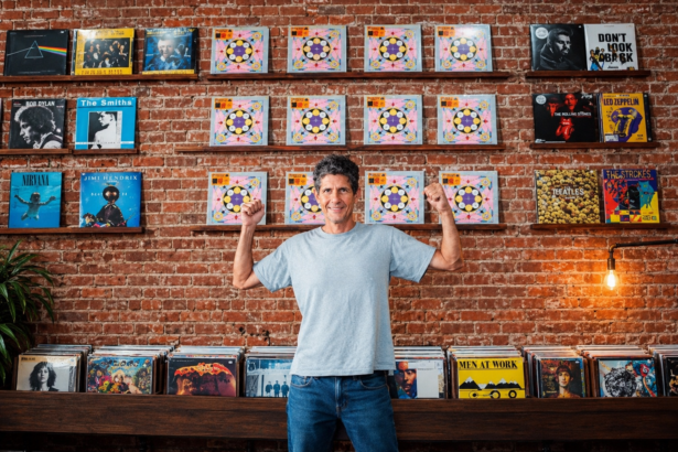 Mike D of the Beastie Boys raises both fists while standing in a record shop, in a light gray T-shirt and jeans before an exposed brick wall lined with vinyl albums.