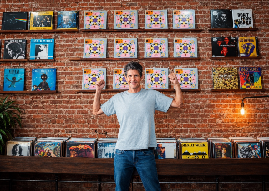 Mike D of the Beastie Boys raises both fists while standing in a record shop, in a light gray T-shirt and jeans before an exposed brick wall lined with vinyl albums.