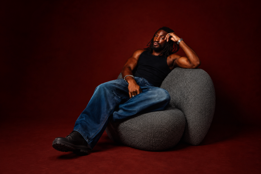 Odeal reclines in a grey pod chair wearing a black tank top and blue jeans against a deep red backdrop, in press art tied to his EP 'For a Good Time.'