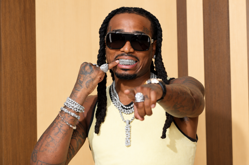 A rapper with long braids points toward the camera, wearing dark sunglasses, a diamond grill, a pale yellow tank top, and multiple diamond chains, rings, and bracelets.