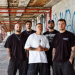 The five members of Pain Of Truth stand together in a graffiti-covered urban walkway with red scaffolding, wearing dark streetwear and chains, their expressions serious.