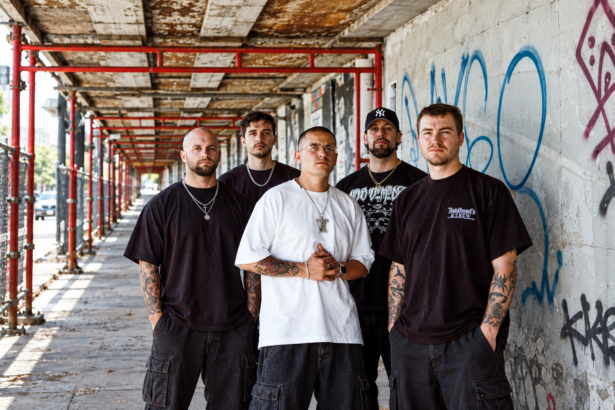 The five members of Pain Of Truth stand together in a graffiti-covered urban walkway with red scaffolding, wearing dark streetwear and chains, their expressions serious.