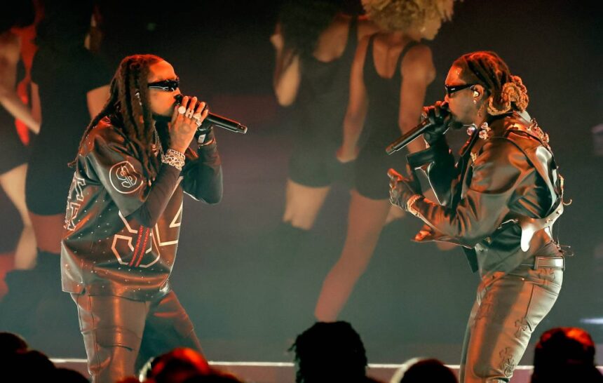 Quavo and Offset of Migos perform together on stage in black leather outfits and sunglasses, each rapping into a microphone under warm red stage lighting.