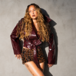 SAM SHI posing against a grey wall in a burgundy croc-effect leather jacket and leopard print shorts