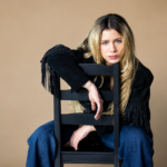Slayyyter sits backwards on a black wooden chair against a tan backdrop, resting her arms over the chair back, wearing a black fringed suede jacket and blue jeans with long blonde hair.