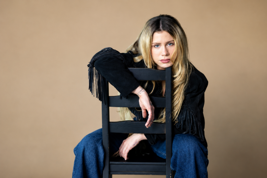Slayyyter sits backwards on a black wooden chair against a tan backdrop, resting her arms over the chair back, wearing a black fringed suede jacket and blue jeans with long blonde hair.