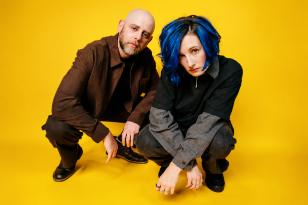 Slothrust duo Leah Wellbaum, with blue hair, and Will Gorin crouch together against a bright yellow background in dark layered clothing, looking toward the camera.