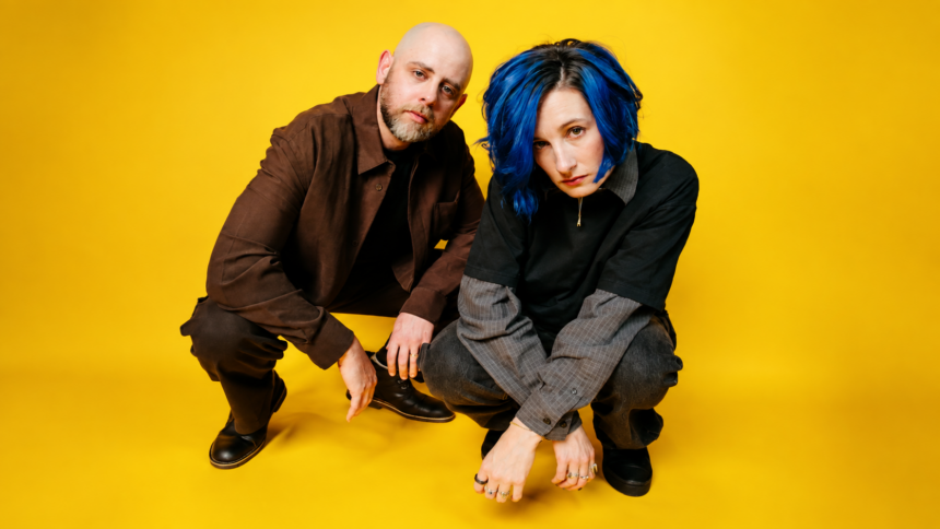 Slothrust duo Leah Wellbaum, with blue hair, and Will Gorin crouch together against a bright yellow background in dark layered clothing, looking toward the camera.