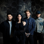 Spiritbox pose in front of a dark hexagonal-patterned backdrop, frontwoman Courtney LaPlante with long dark hair in black beside her three bandmates in denim and dark jackets.