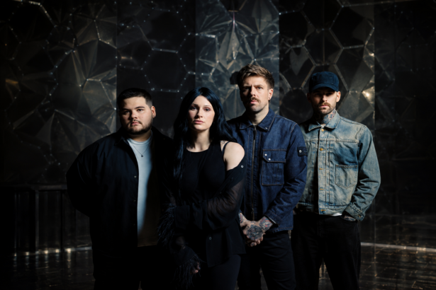Spiritbox pose in front of a dark hexagonal-patterned backdrop, frontwoman Courtney LaPlante with long dark hair in black beside her three bandmates in denim and dark jackets.