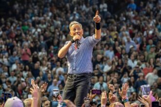 Bruce Springsteen points skyward while singing into a microphone on a stage surrounded by a packed arena crowd with raised hands and phones, during his Land of Hope and Dreams tour.