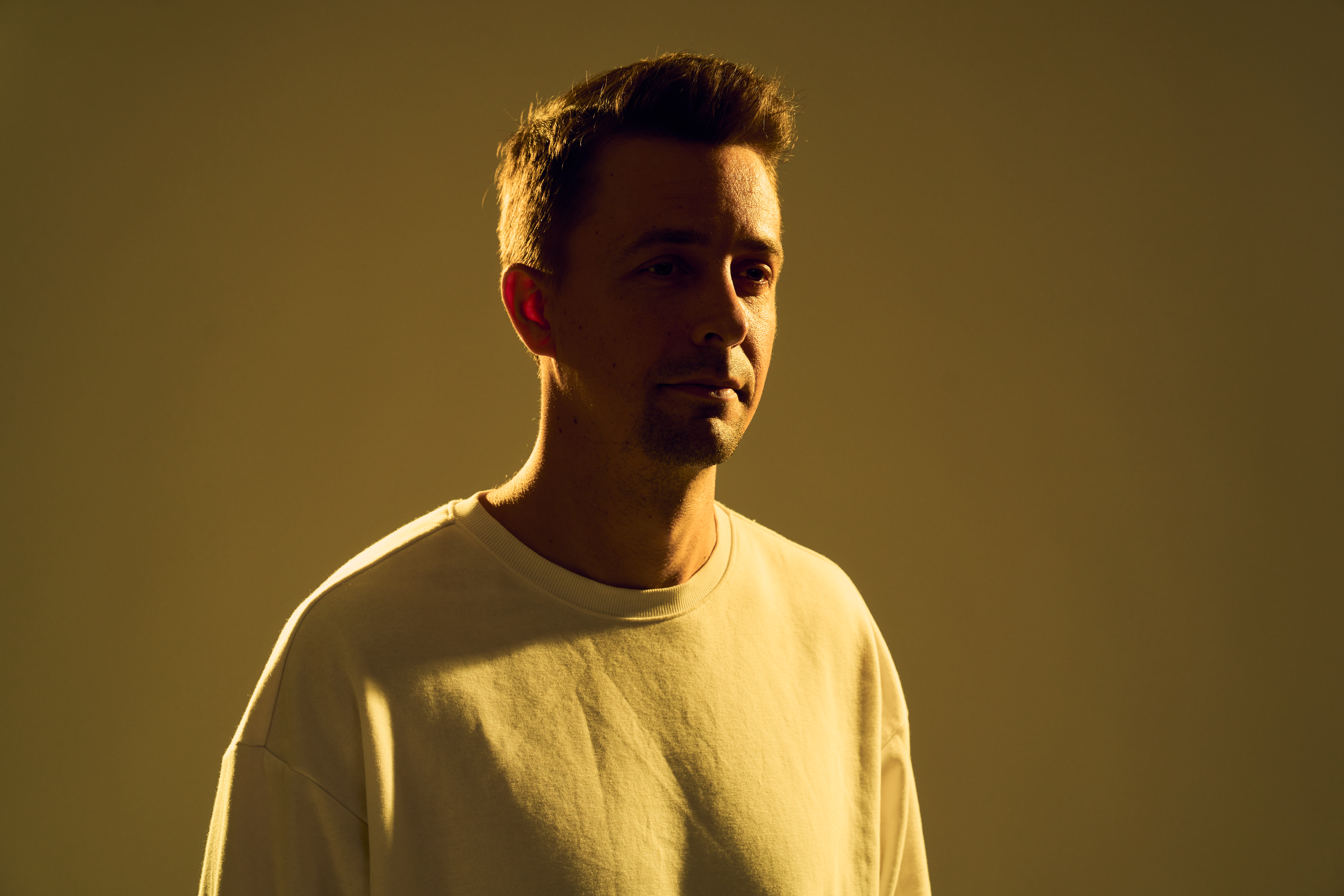 Steve Stinson stands in a cream sweatshirt under warm golden lighting against a muted backdrop, in press art for his single 'Evergreen.'
