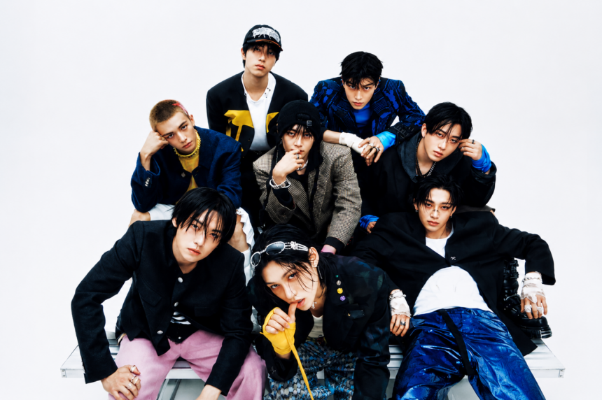 The eight members of Stray Kids pose together against a white backdrop in edgy streetwear and tailored jackets, arranged in a layered group formation with cool, direct expressions.