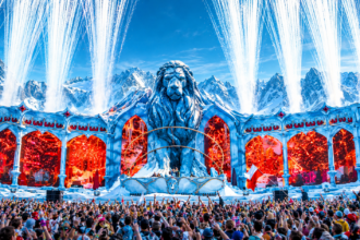 Tomorrowland Winter 2027 poster showing the festival's lion mainstage against snowy mountains with pyrotechnics and a packed crowd, captioned March 20 to 27, 2027, Alpe d'Huez, France.