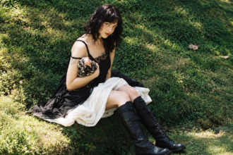 Violet Grohl sits on a grassy hillside in a black slip dress with lace detailing and knee-high lace-up leather boots, her dark wavy hair loose as she gazes toward the camera in dappled sunlight.