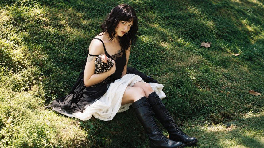 Violet Grohl sits on a grassy hillside in a black slip dress with lace detailing and knee-high lace-up leather boots, her dark wavy hair loose as she gazes toward the camera in dappled sunlight.