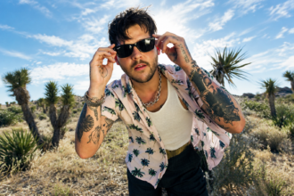 Nathan Williams of Wavves adjusts his sunglasses in a desert landscape, wearing an open palm-print shirt over a white tank top with a silver chain and visible arm tattoos.