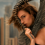 Cara Delevingne pulls a black and cream striped knit over her head and across her face, her tousled hair blowing, with a blurred city skyline behind her.