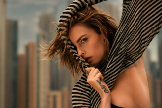 Cara Delevingne pulls a black and cream striped knit over her head and across her face, her tousled hair blowing, with a blurred city skyline behind her.