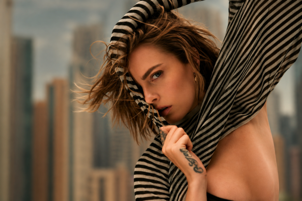 Cara Delevingne pulls a black and cream striped knit over her head and across her face, her tousled hair blowing, with a blurred city skyline behind her.