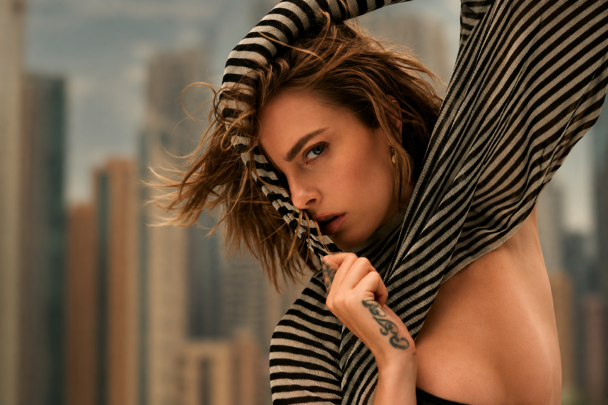 Cara Delevingne pulls a black and cream striped knit over her head and across her face, her tousled hair blowing, with a blurred city skyline behind her.