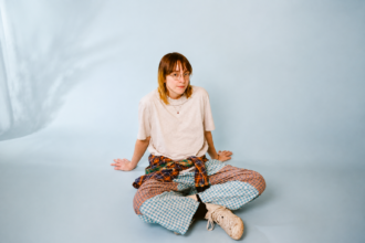 Emily A. Sprague sits cross-legged on the floor of a pale blue studio, wearing wire-rimmed glasses, a cream T-shirt, a plaid shirt tied at the waist and patchwork plaid trousers.