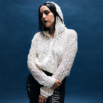 Logic1000 stands against a deep blue backdrop in a white textured hooded top, dark lipstick and slicked hair, arms crossed and eyes lowered.
