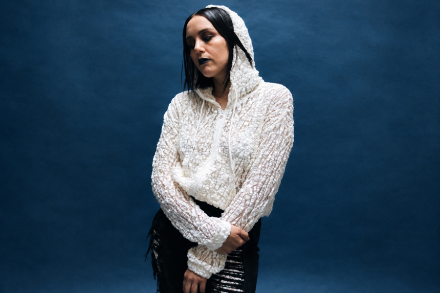 Logic1000 stands against a deep blue backdrop in a white textured hooded top, dark lipstick and slicked hair, arms crossed and eyes lowered.