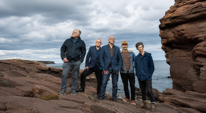 The five members of Teenage Fanclub stand on red sandstone rocks by a grey, overcast sea, in jackets and blazers, looking off in different directions.