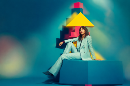 Anna Meredith, in a pale mint suit, sits on a large teal block in front of a stack of pink, yellow and teal geometric shapes against a blurred teal backdrop.
