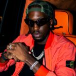 Ayjay Bobo in cat-eye sunglasses, a backward green trucker cap and an orange bomber jacket, seated in a car with orange leather seats.