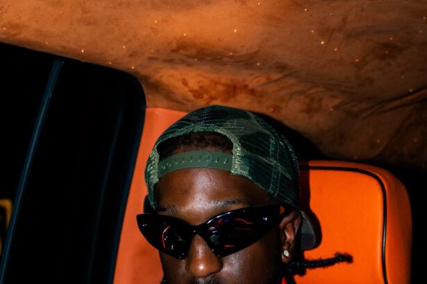 Ayjay Bobo in cat-eye sunglasses, a backward green trucker cap and an orange bomber jacket, seated in a car with orange leather seats.