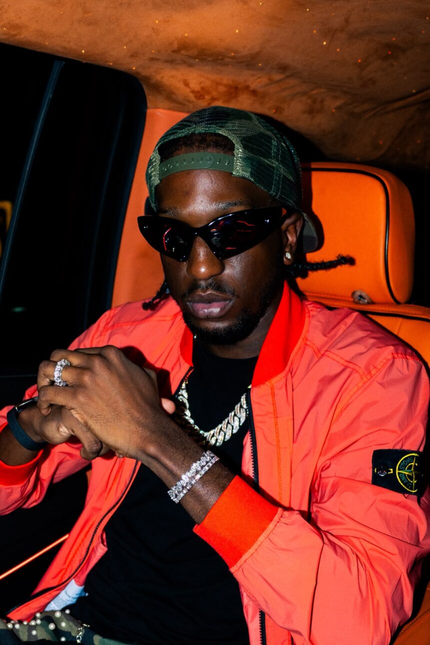 Ayjay Bobo in cat-eye sunglasses, a backward green trucker cap and an orange bomber jacket, seated in a car with orange leather seats.