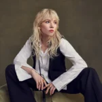 Carly Rae Jepsen crouches barefoot on draped olive fabric, wearing an oversized white shirt, black waistcoat and black trousers, with wavy blonde hair and a fringe against an olive backdrop.
