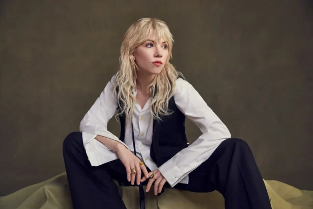 Carly Rae Jepsen crouches barefoot on draped olive fabric, wearing an oversized white shirt, black waistcoat and black trousers, with wavy blonde hair and a fringe against an olive backdrop.