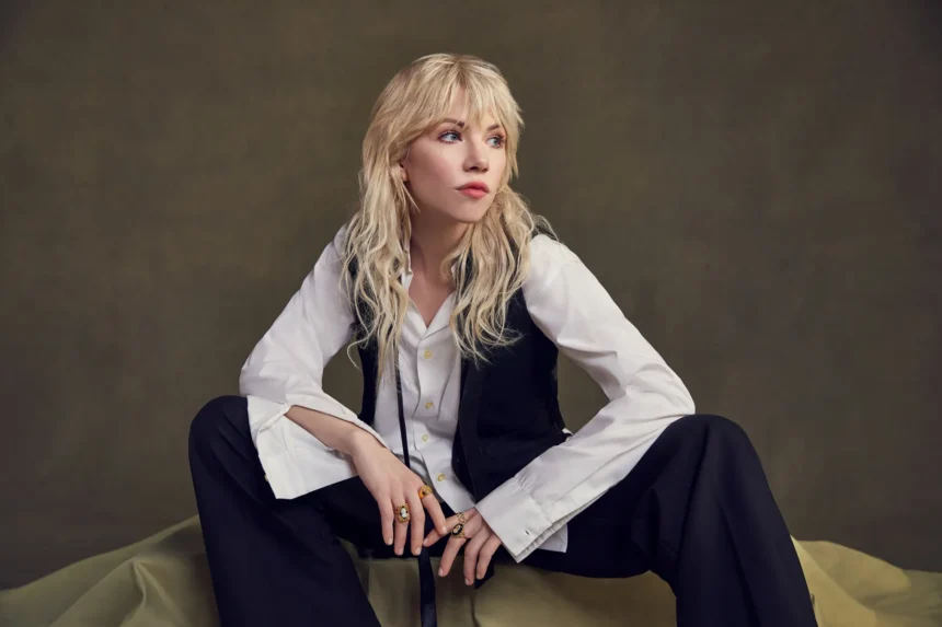 Carly Rae Jepsen crouches barefoot on draped olive fabric, wearing an oversized white shirt, black waistcoat and black trousers, with wavy blonde hair and a fringe against an olive backdrop.