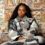Cristale sits in a grey and cream patchwork monogram denim set, with long braids, against ornate hand-painted floral wallpaper.